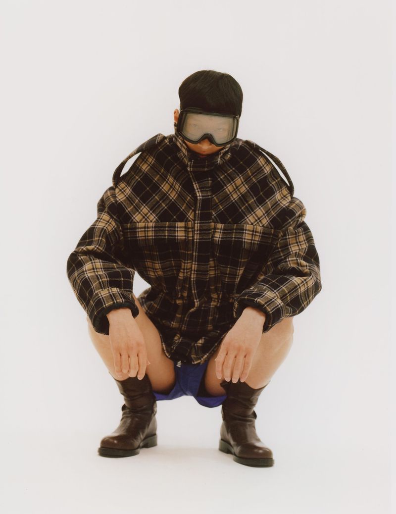 Man squats in photo studio wearing cap, goggles, plaid jacket, blue shorts, and brown boots against a solid white backdrop.