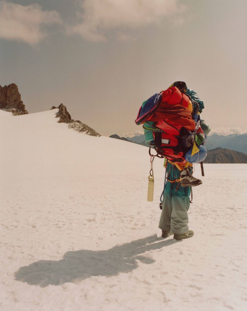 Person heavily loaded with gear ascends snowy, mountainous terrain under a cloudy sky, casting a long shadow on the snowfield.