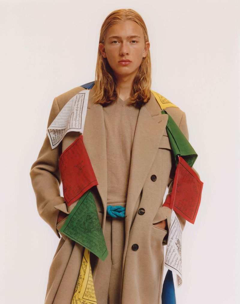 Adult male fashion model poses indoors wearing a beige overcoat with colorful prayer flags draped around the shoulders against a white backdrop.
