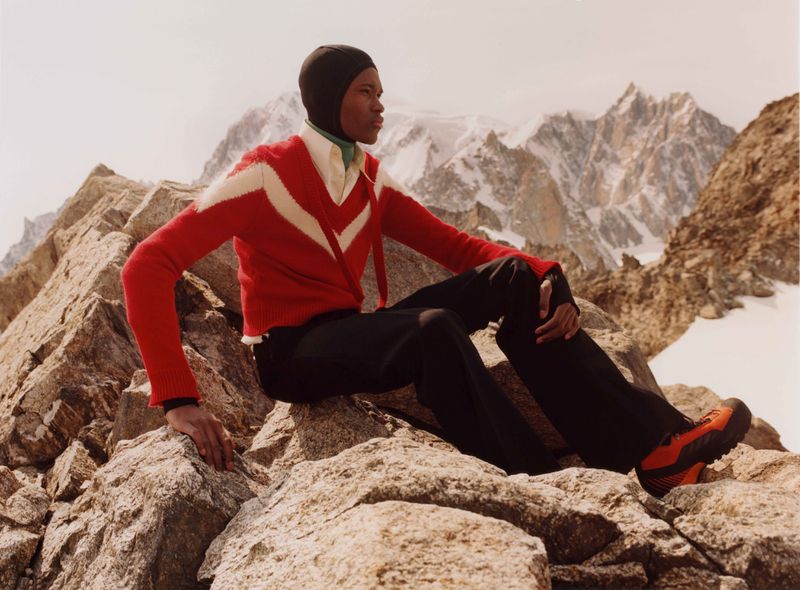 An adult poses outdoors in a ski mask, red cardigan, black pants, and orange hiking boots, seated on rocks with mountains in background.