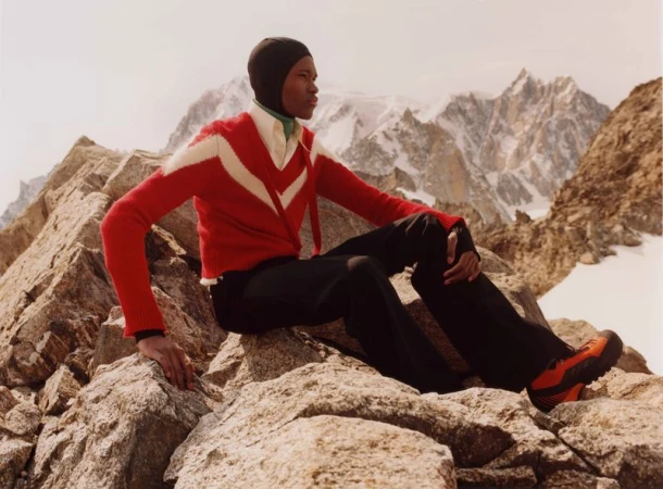 An adult poses outdoors in a ski mask, red cardigan, black pants, and orange hiking boots, seated on rocks with mountains in background.