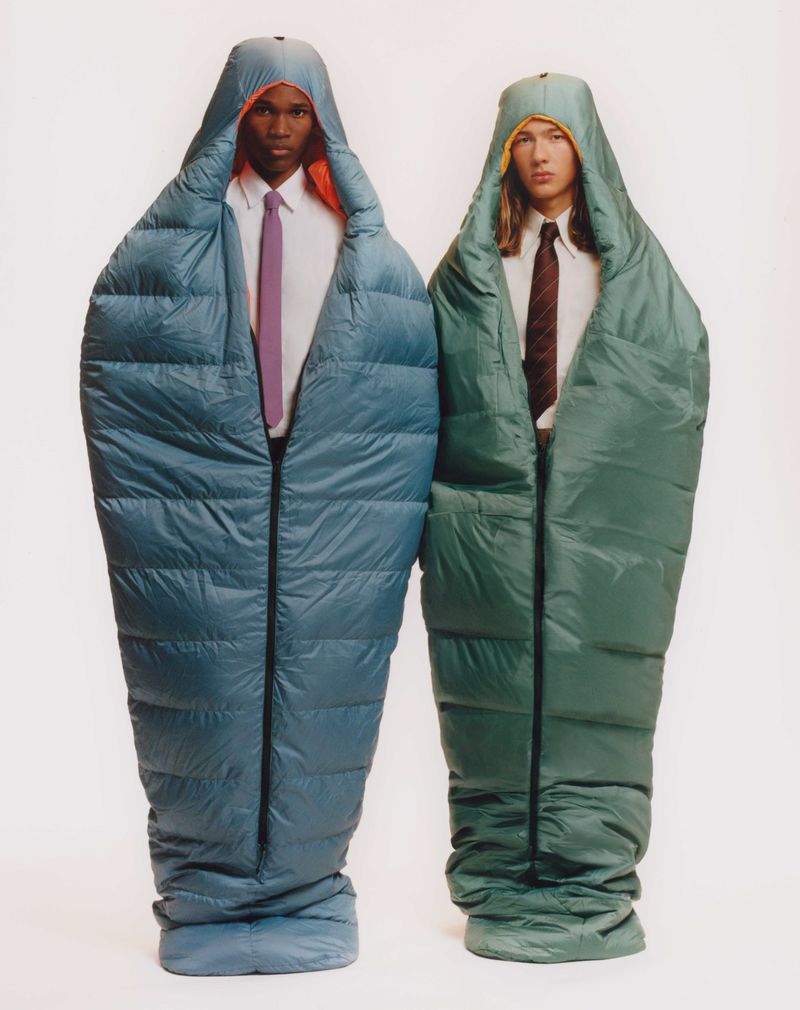 Two men in teal and green sleeping bags standing side-by-side in front of a plain white background, wearing ties and collared shirts.