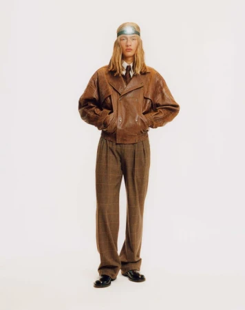 Model with long blonde hair wearing a brown leather jacket and plaid trousers against a plain white background