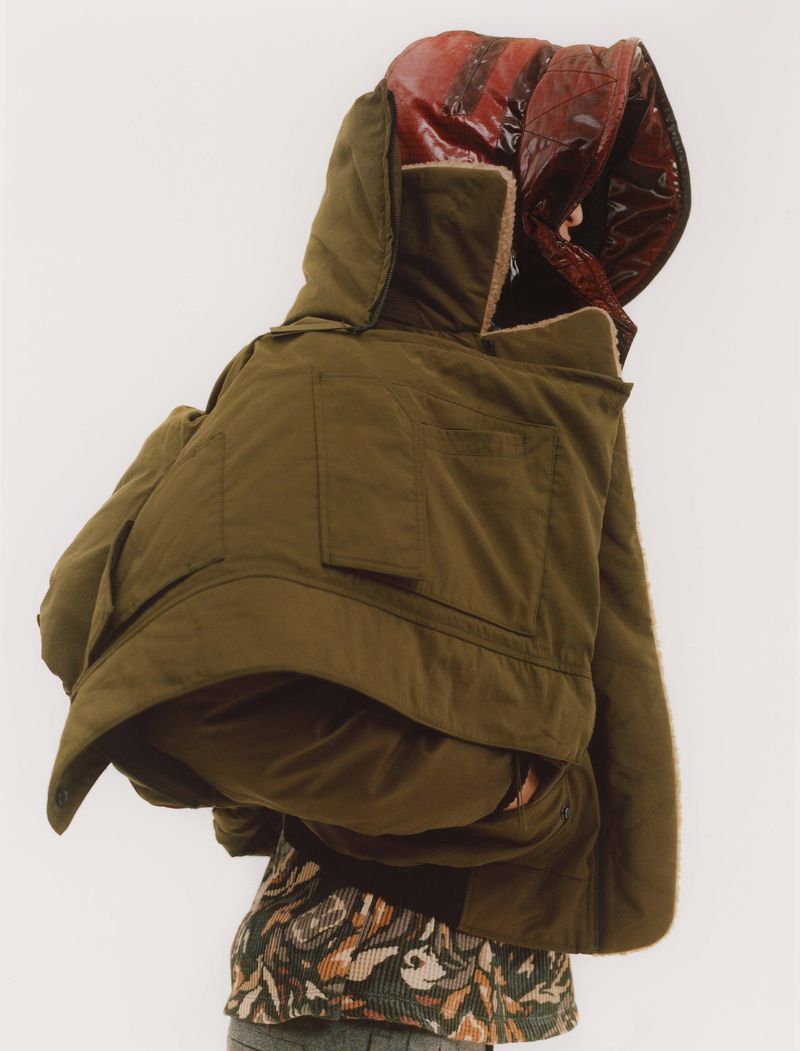 Model wearing an oversized olive green hooded jacket over a patterned top against a plain white background