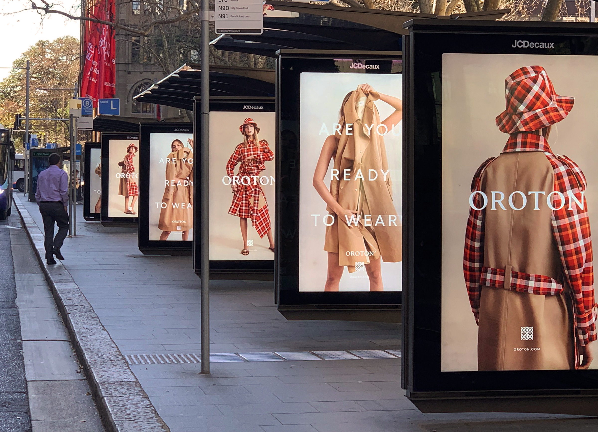 Outdoor bus stop with fashion advertising posters featuring women's tan and red-plaid overcoats, dresses, and hats by Oroton.