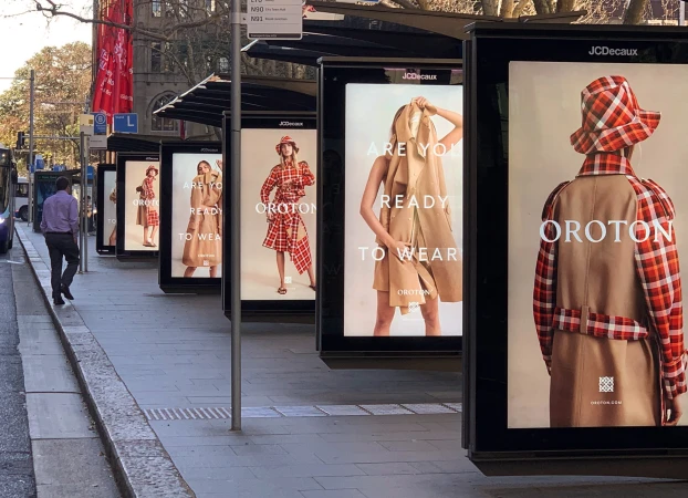 Outdoor bus stop with fashion advertising posters featuring women's tan and red-plaid overcoats, dresses, and hats by Oroton.