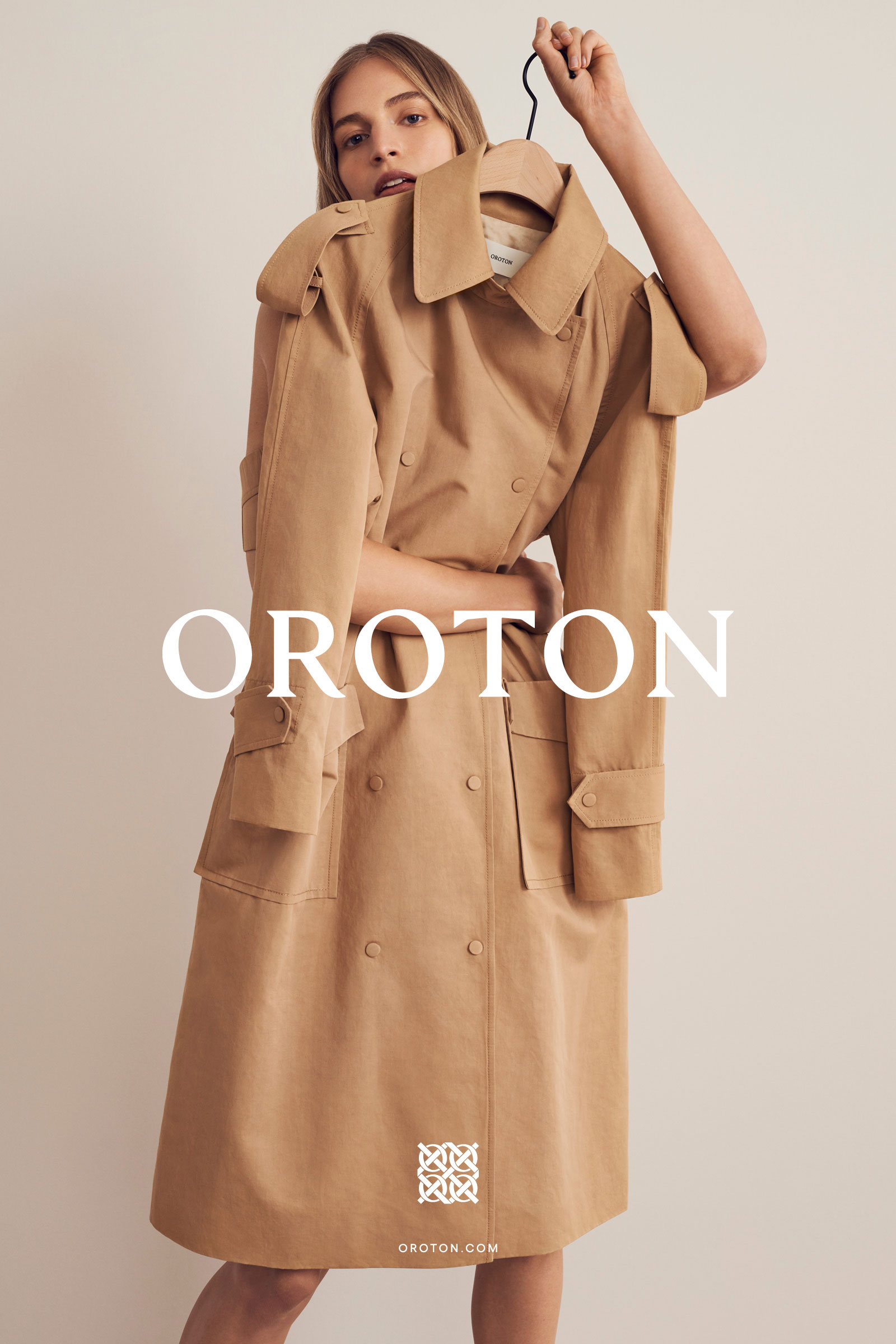 Model holding a tan trench coat up in front of her face against a plain light background