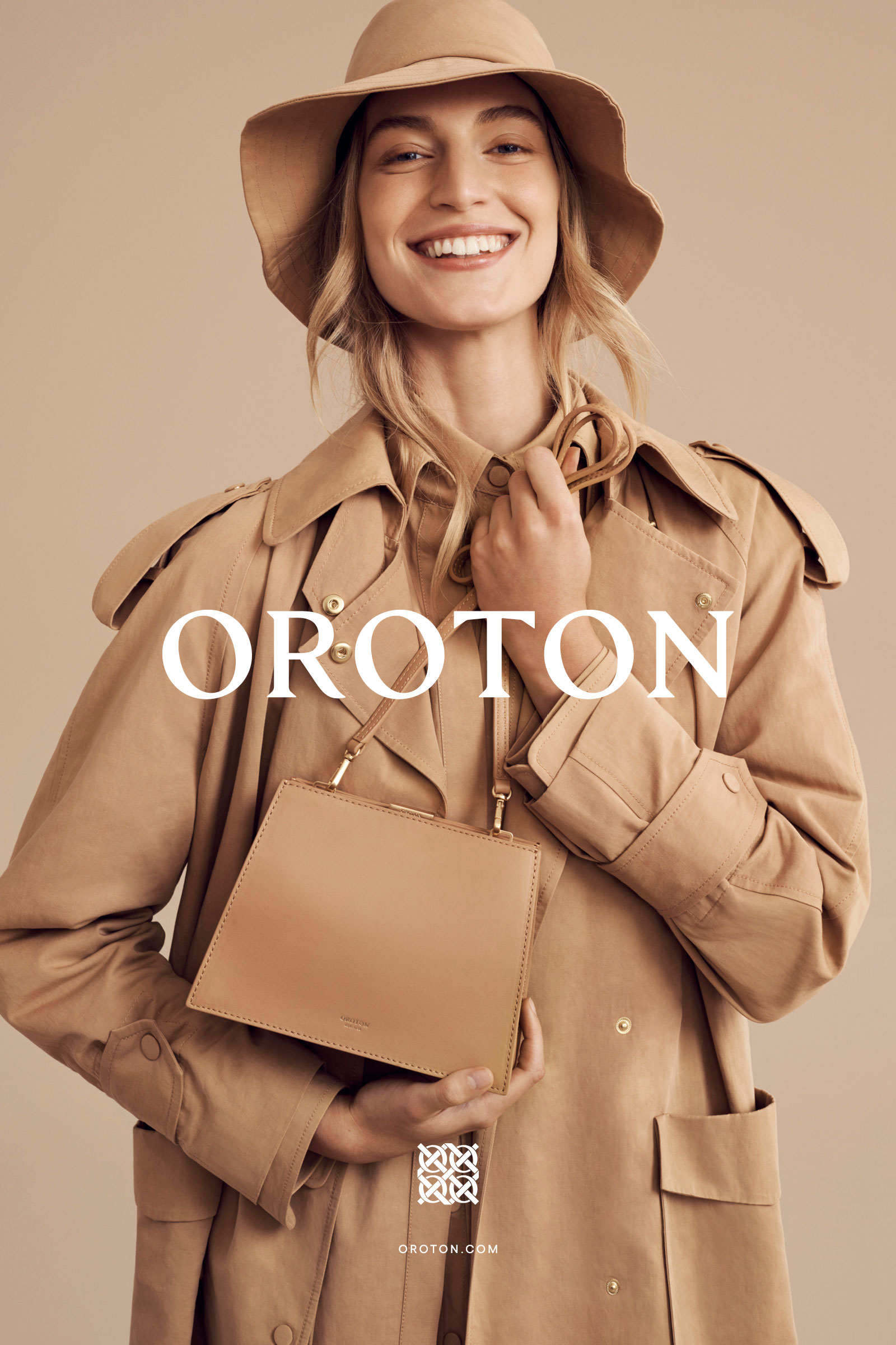 Smiling fashion model in a beige trench coat and hat poses with a small, square beige handbag against a matching monochromatic backdrop.