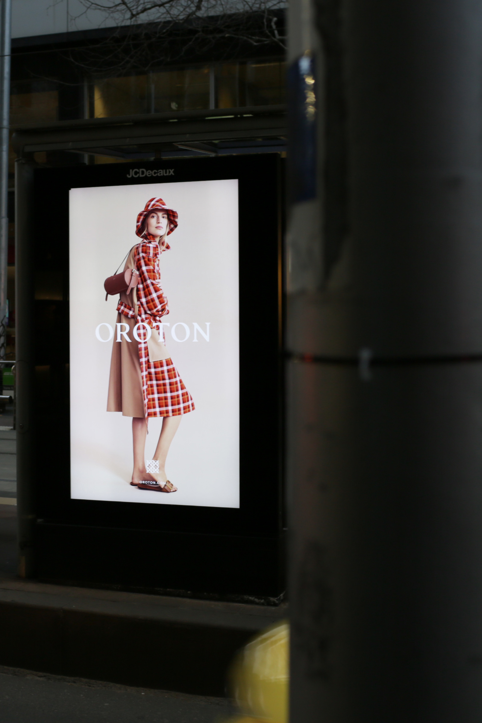 Outdoor advertising billboard featuring a stylish woman in a red plaid outfit and hat for Oroton, showcasing fashion and urban advertising.