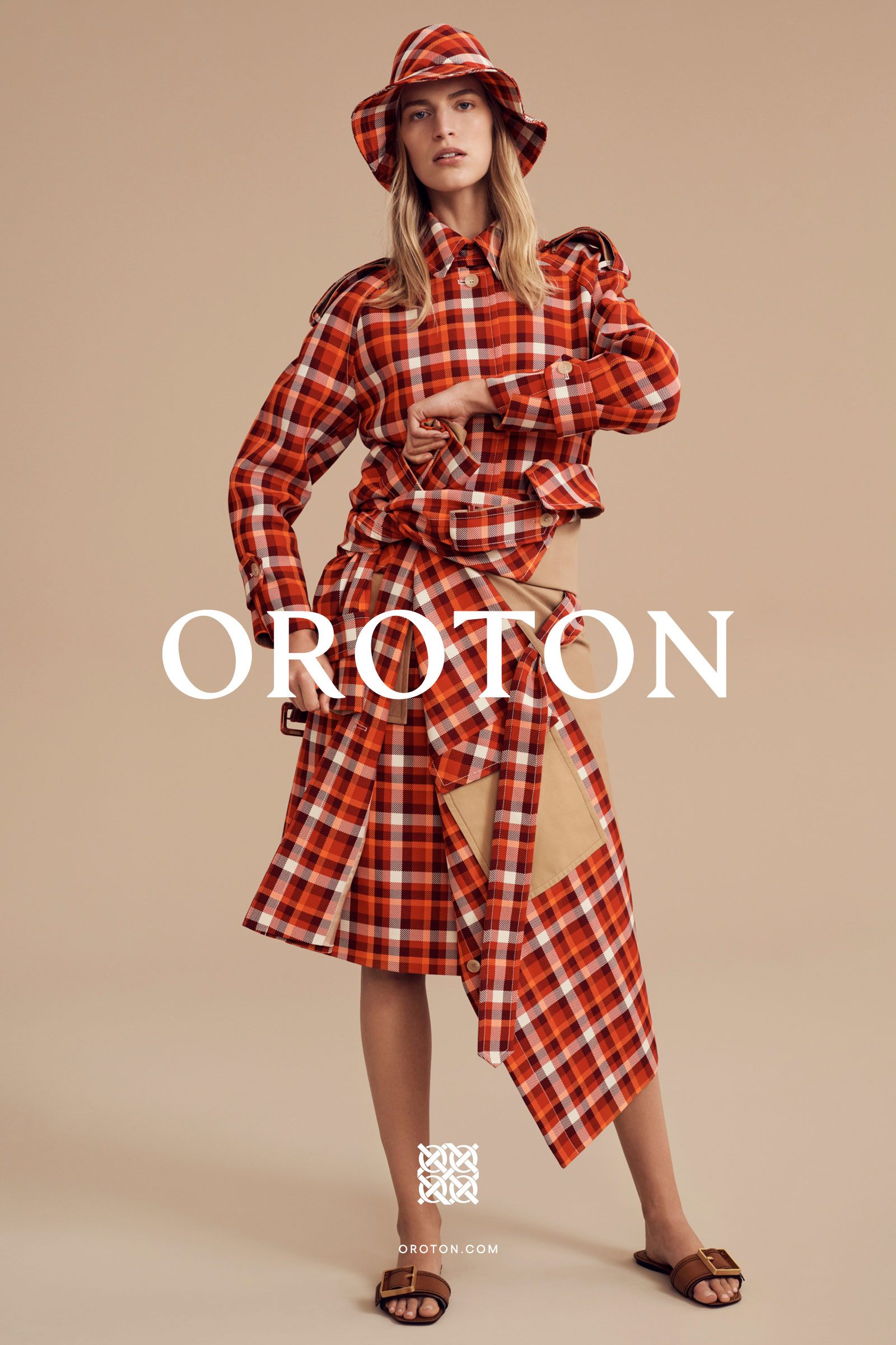 A woman in a red and white plaid outfit with a matching hat poses in a studio against a tan background for a fashion shot.