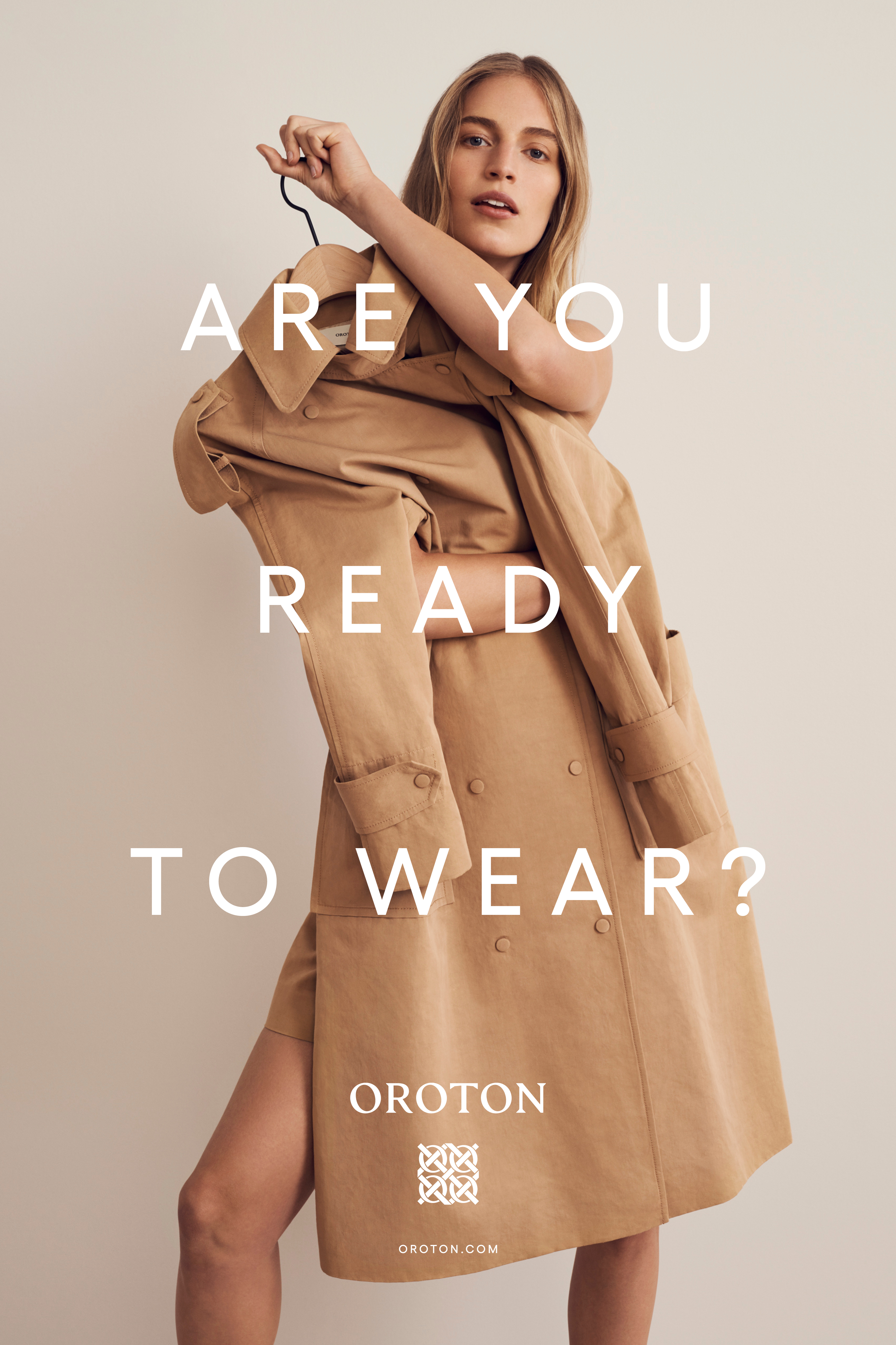 Fashion shot of a female model holding a tan trench coat on a hanger, standing indoors against a neutral background, promoting Oroton.
