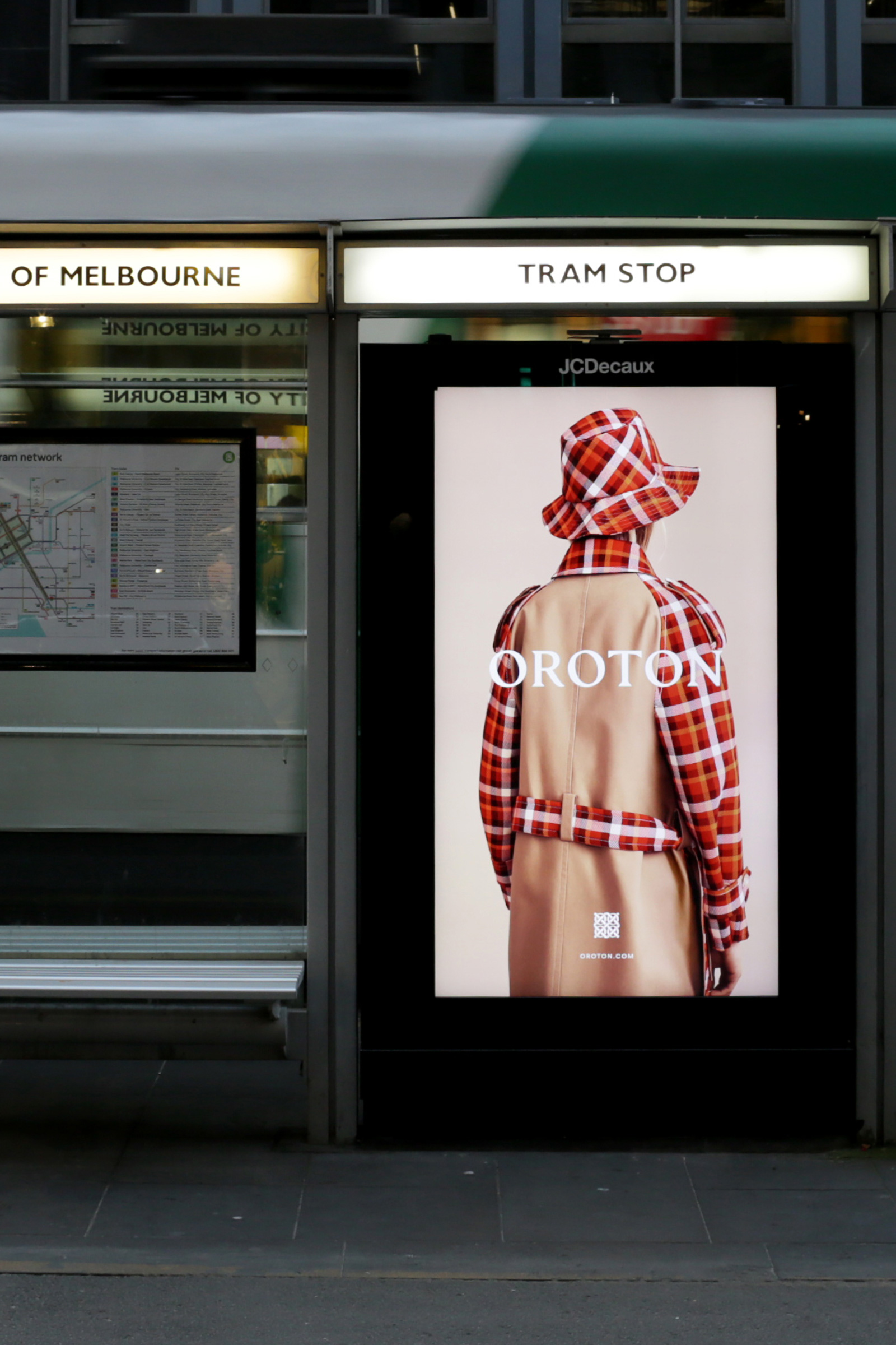 Fashion advertisement at a tram stop featuring a person wearing a plaid hat and coat with a tan trench coat over the top.