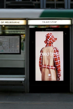 Fashion advertisement at a tram stop featuring a person wearing a plaid hat and coat with a tan trench coat over the top.
