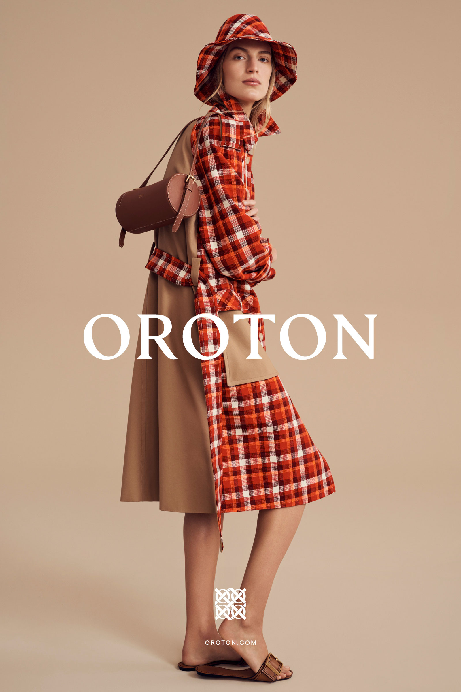 Model wearing a plaid shirt dress, trench coat, bucket hat, and carrying a leather cylindrical shoulder bag.