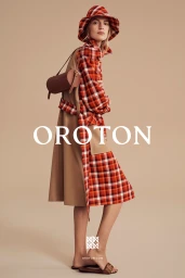 Model wearing red plaid bucket hat and shirt with a beige trench coat carrying a brown cylindrical handbag