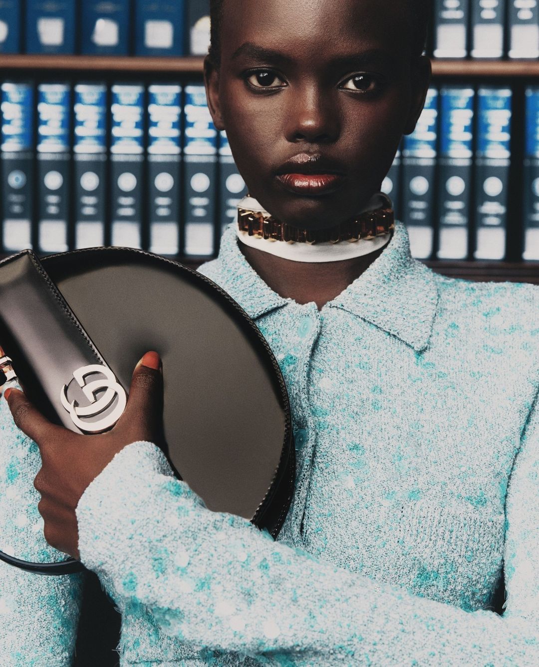 Close-up of an African-American woman in a button-down, holding a Gucci handbag, against a bookshelf background in a fashion editorial.