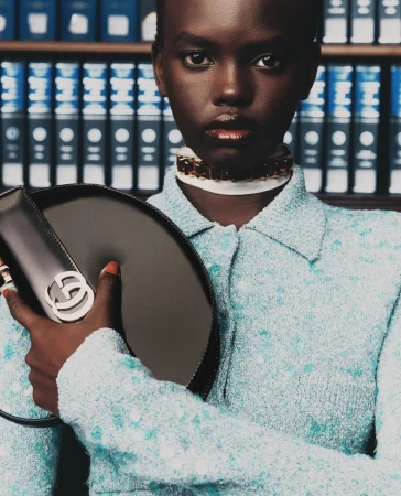 Close-up of an African-American woman in a button-down, holding a Gucci handbag, against a bookshelf background in a fashion editorial.