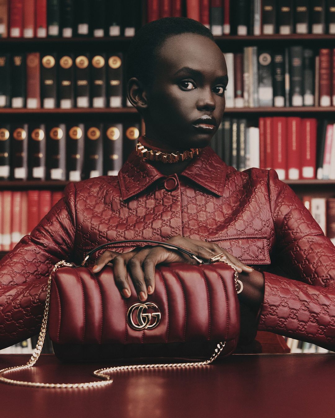 A portrait of a woman in a red Gucci jacket with a red Gucci purse in front of a bookcase in an indoor environment.
