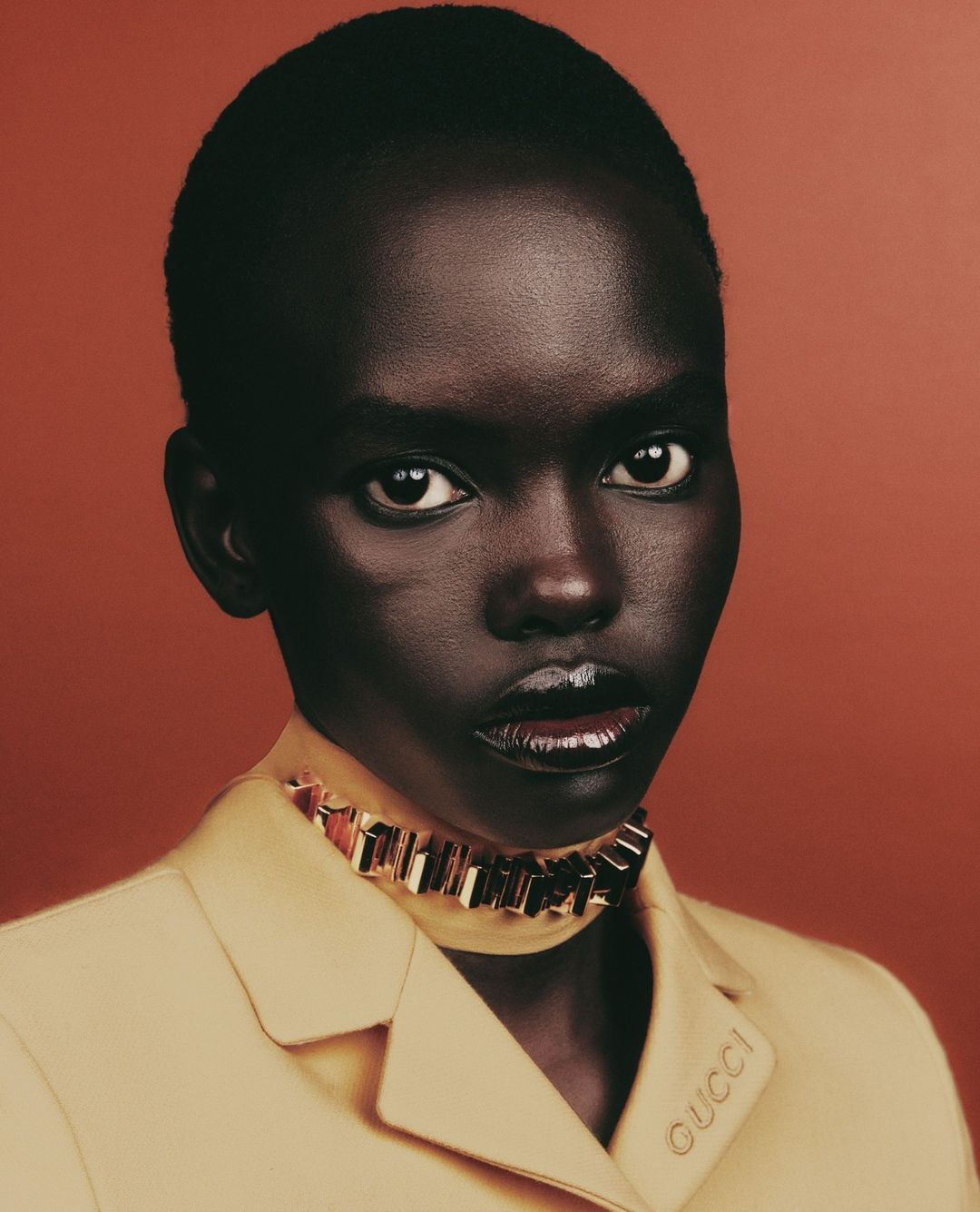 Portrait of an African-American woman with dark skin wearing a yellow blazer and jewelry against a solid reddish-brown background.