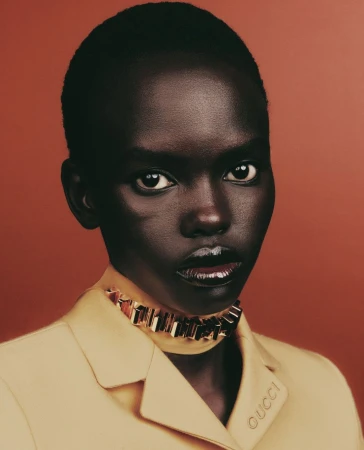 Portrait of an African-American woman with dark skin wearing a yellow blazer and jewelry against a solid reddish-brown background.