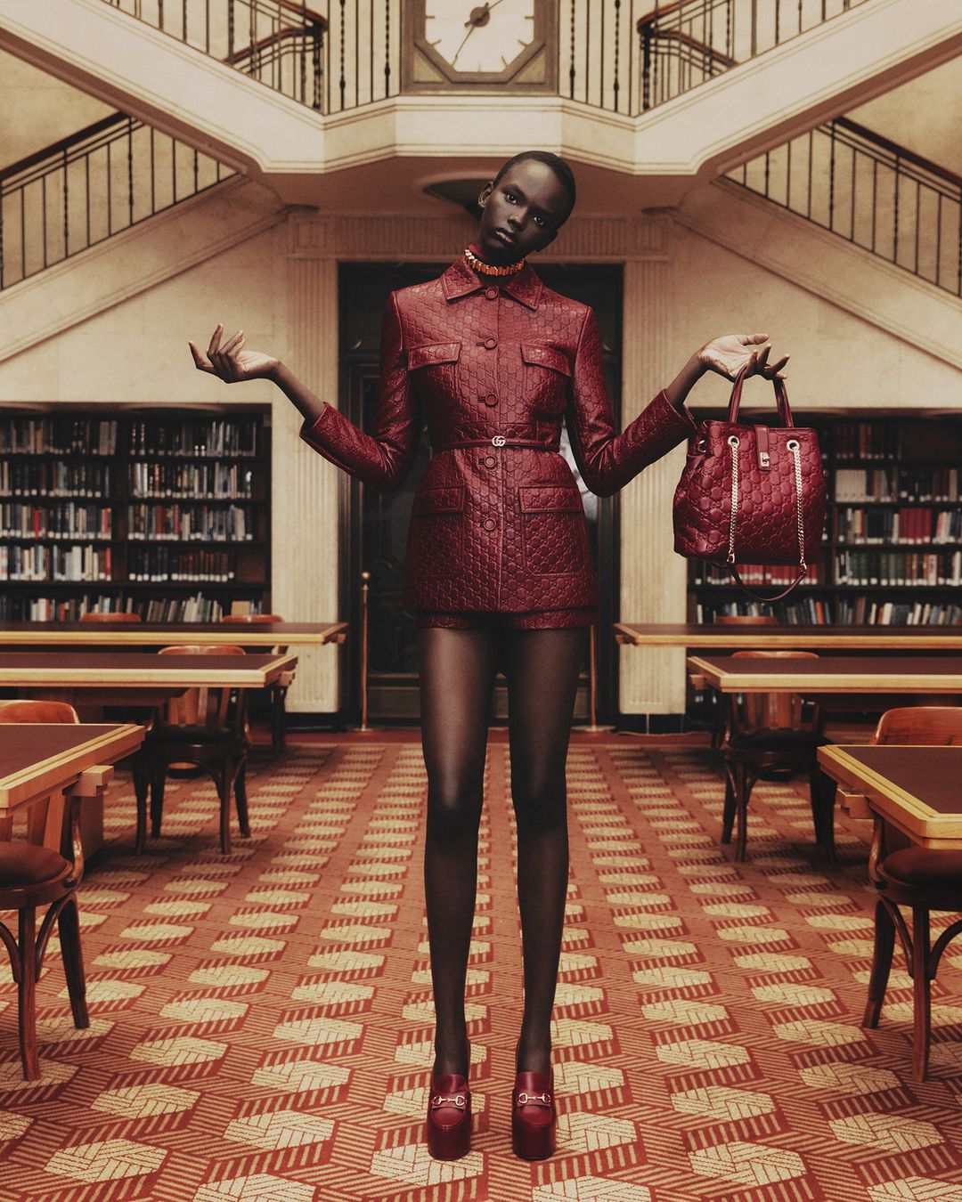 Fashion shot of a dark-skinned woman in a red suit, posing in a library, holding a matching purse, and wearing red platform heels.