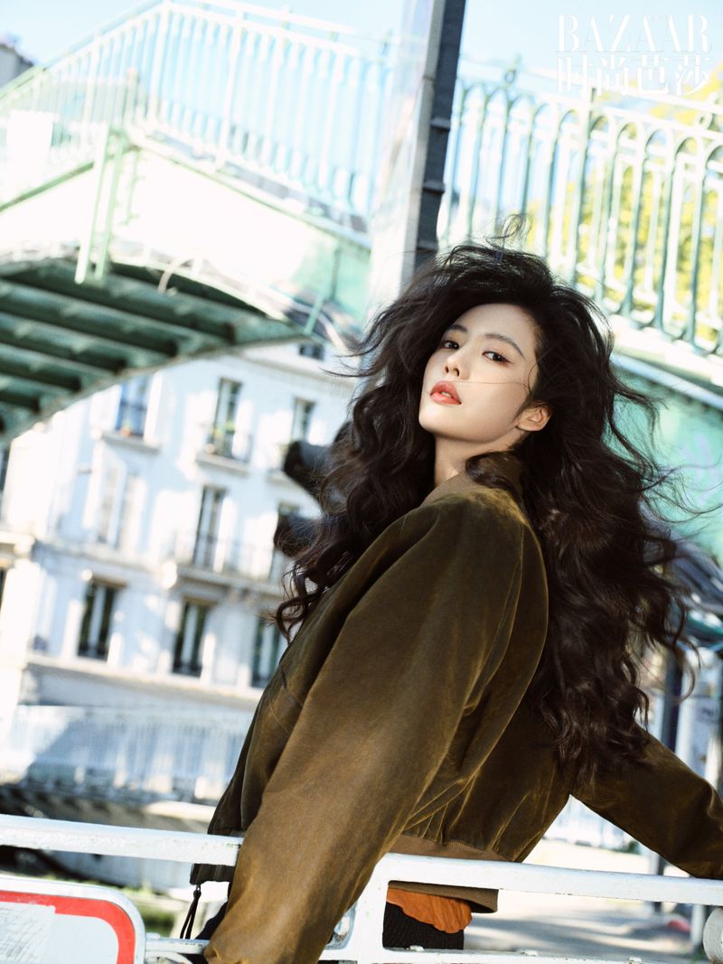 Portrait of a woman with long dark hair and olive jacket posing in front of buildings and a bridge on a sunny day.