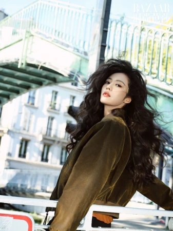 Portrait of a woman with long dark hair and olive jacket posing in front of buildings and a bridge on a sunny day.