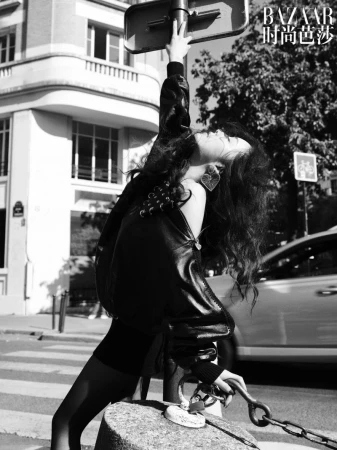 Black and white fashion shot of a woman in a jacket, dress, and jewelry leaning against a traffic sign pole on a city street.