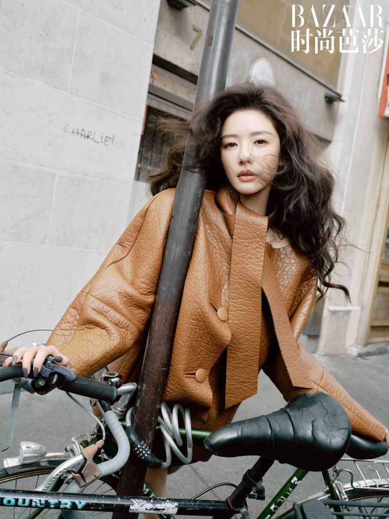 A stylish woman with dark hair poses with a bicycle in an urban setting, wearing a tan jacket for a fashion editorial.