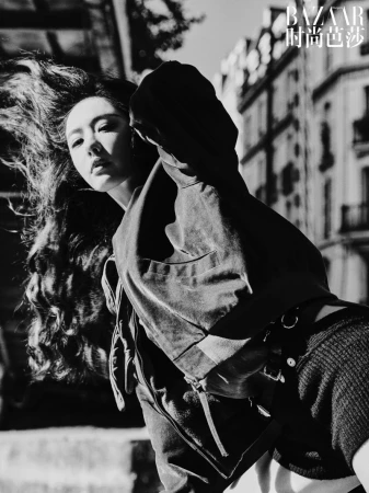 Black and white editorial fashion portrait of a woman with long dark hair blowing in the wind against an urban street background.