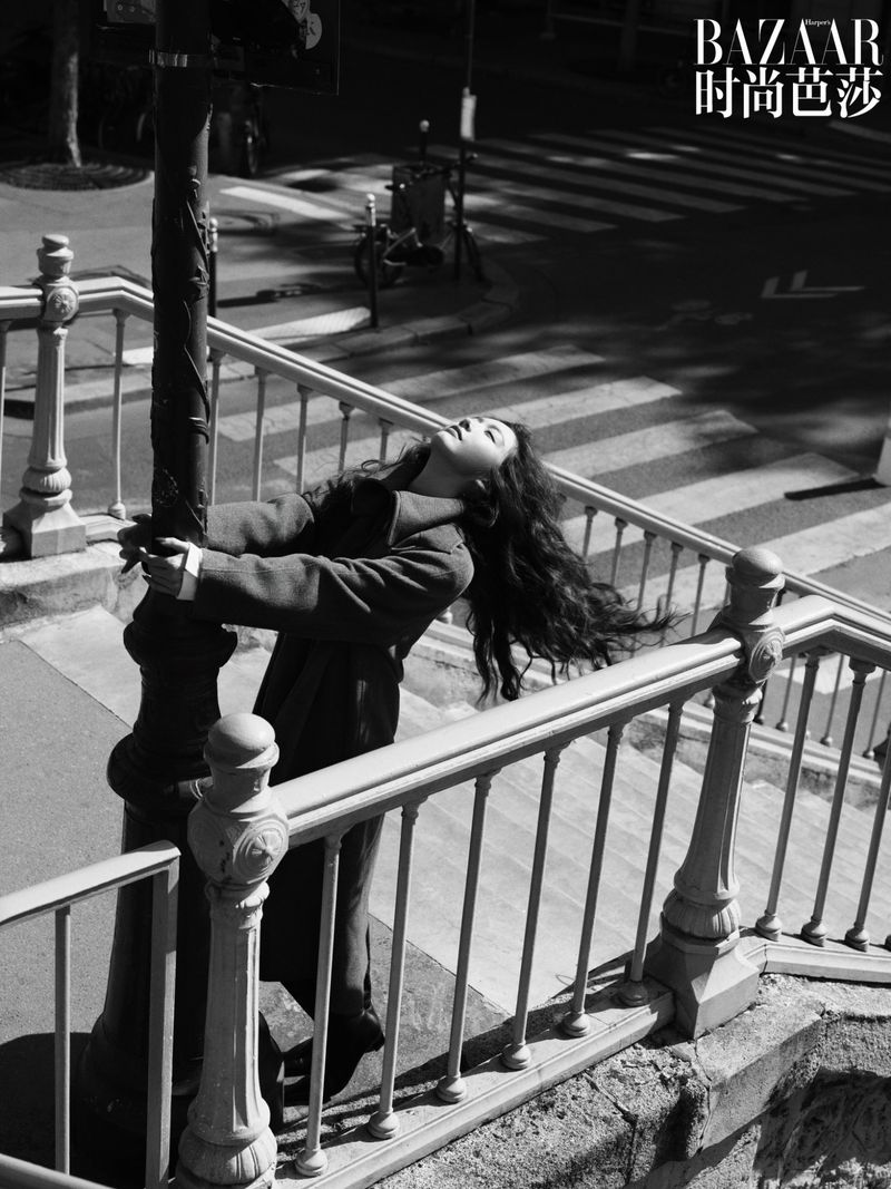 Monochrome fashion shot: A woman in a coat leans back against a lamppost, her head tilted back, on an urban staircase with railings.