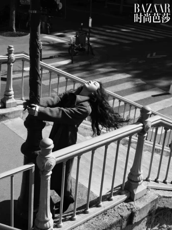 Monochrome fashion shot: A woman in a coat leans back against a lamppost, her head tilted back, on an urban staircase with railings.