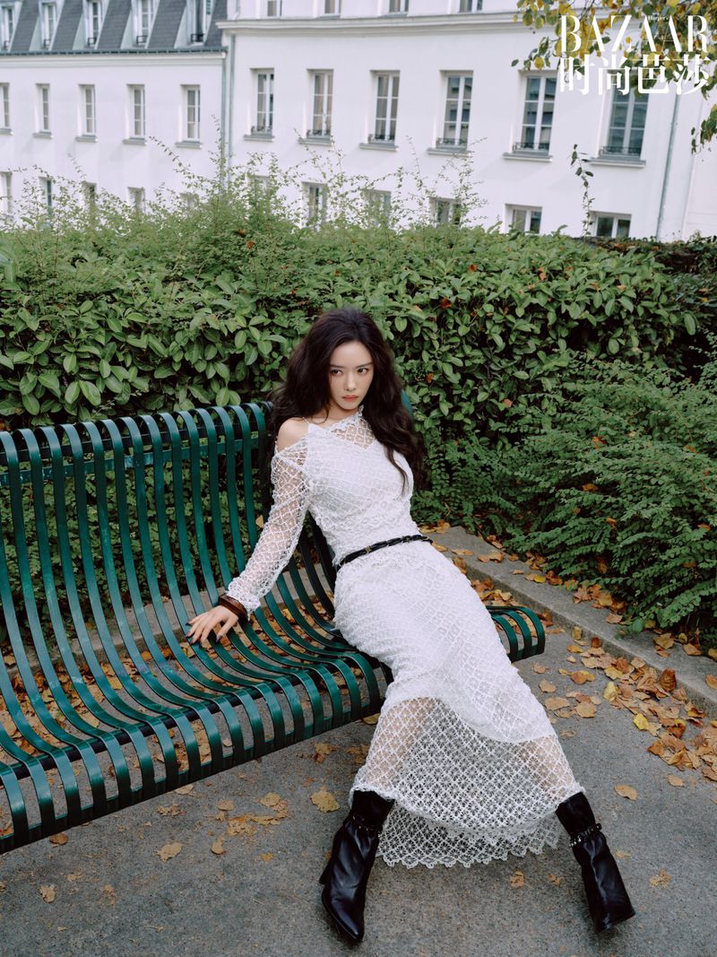 Elegant woman in a long white lace dress and black boots sitting on a park bench with a white building and trees in the background.