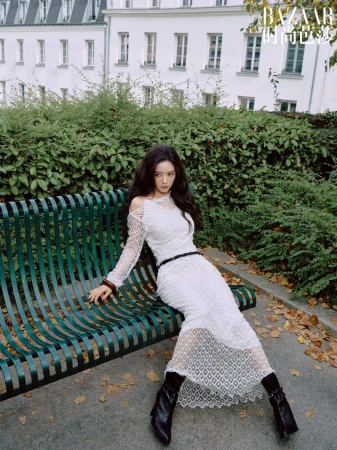 Woman in a long white lace dress sitting on a green park bench in front of a white building with windows.
