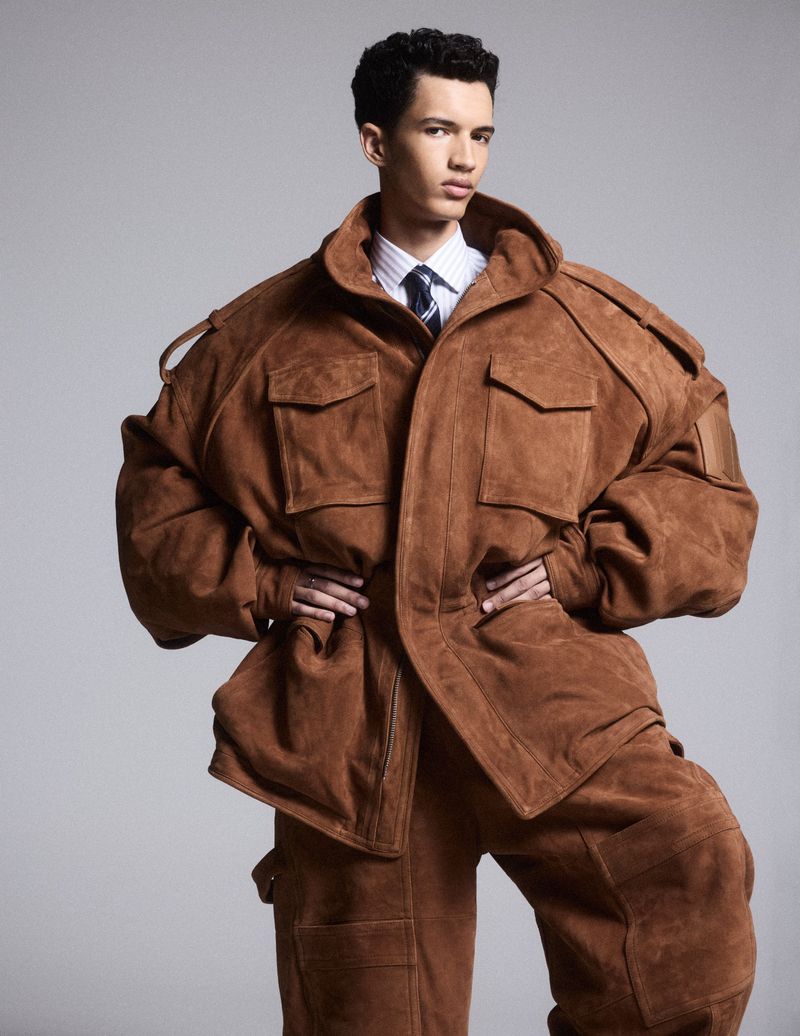 Man in oversized brown suede jacket and matching pants against grey background, posing with hands on hips, in a fashion shot.