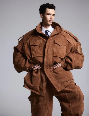 Man in oversized brown suede jacket and matching pants against grey background, posing with hands on hips, in a fashion shot.