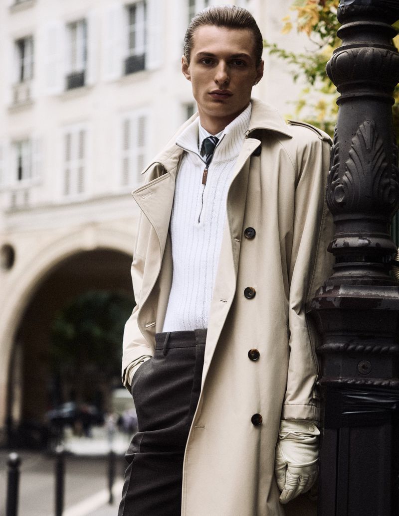 Stylish man in trench coat and gloves stands near lamppost in a European city with historic buildings, captured in soft, natural light.
