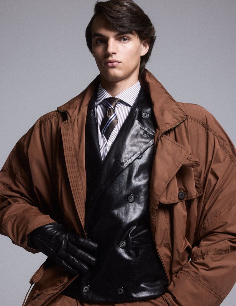 Young man poses in brown trench coat and black vest against a grey background, showcasing a fashion-forward and confident style.