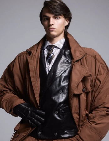 Young man poses in brown trench coat and black vest against a grey background, showcasing a fashion-forward and confident style.
