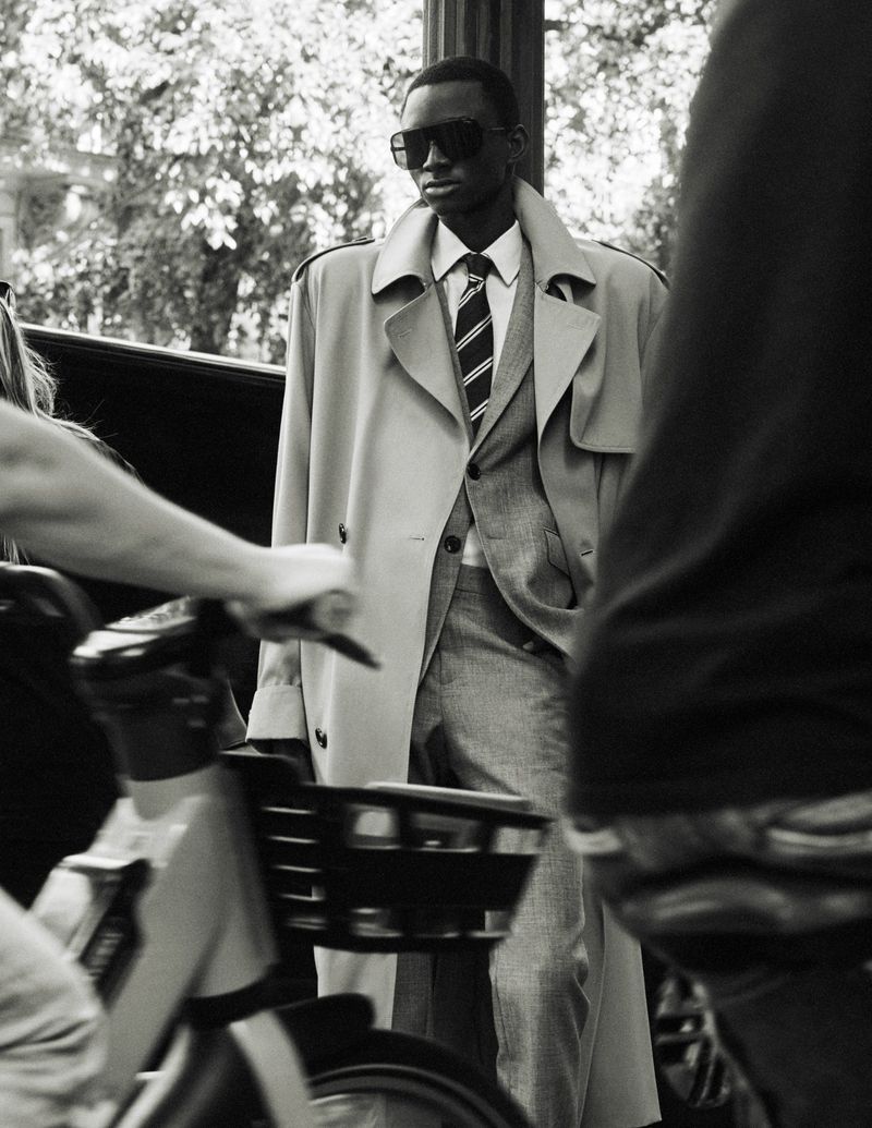 Model in sunglasses and trench coat standing outdoors with a bicycle in the foreground