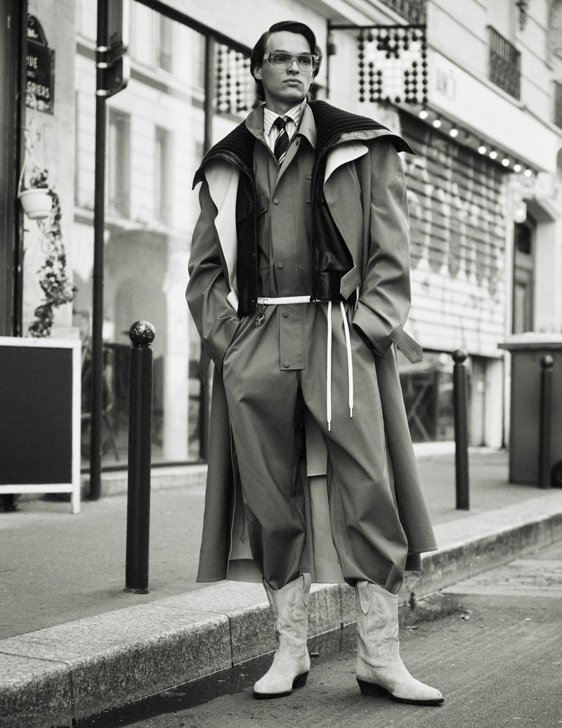 A man in a layered outfit poses on a city street in a black and white shot featuring a coat, jacket, suit, boots, and glasses.