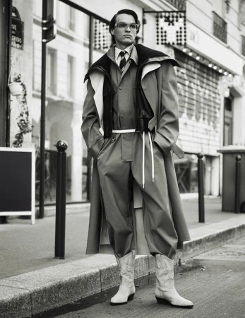 A man in a layered outfit poses on a city street in a black and white shot featuring a coat, jacket, suit, boots, and glasses.