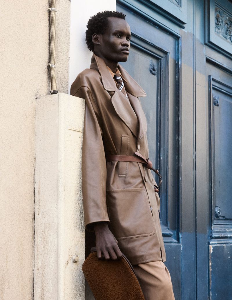 Dark-skinned male model in a brown leather trench coat with brown bag poses against a wall near a blue door in an urban setting.