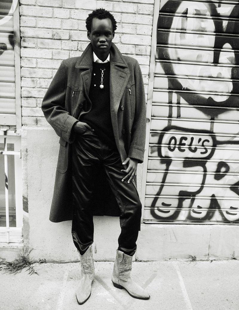 Monochrome portrait of an adult male in an overcoat, sweater, and cowboy boots standing against a graffiti-covered brick wall and metal door.