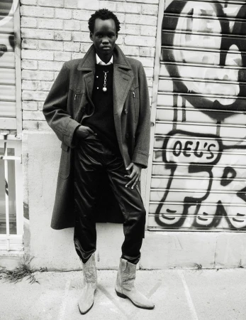 Monochrome portrait of an adult male in an overcoat, sweater, and cowboy boots standing against a graffiti-covered brick wall and metal door.