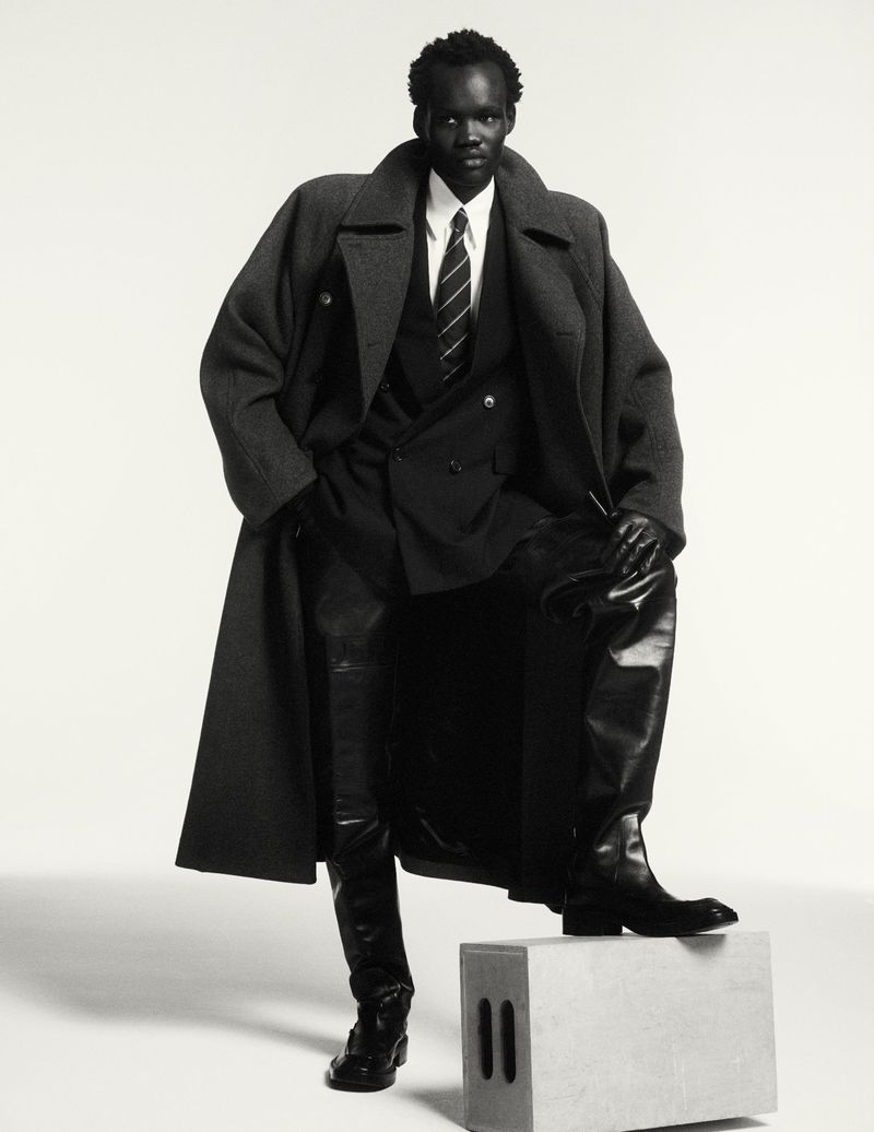 Fashion shot of African male model in wool coat, leather pants, and black boots, striking a pose on a grey cube in studio.