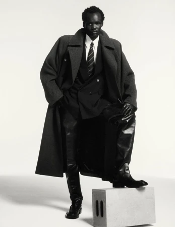 Fashion shot of African male model in wool coat, leather pants, and black boots, striking a pose on a grey cube in studio.