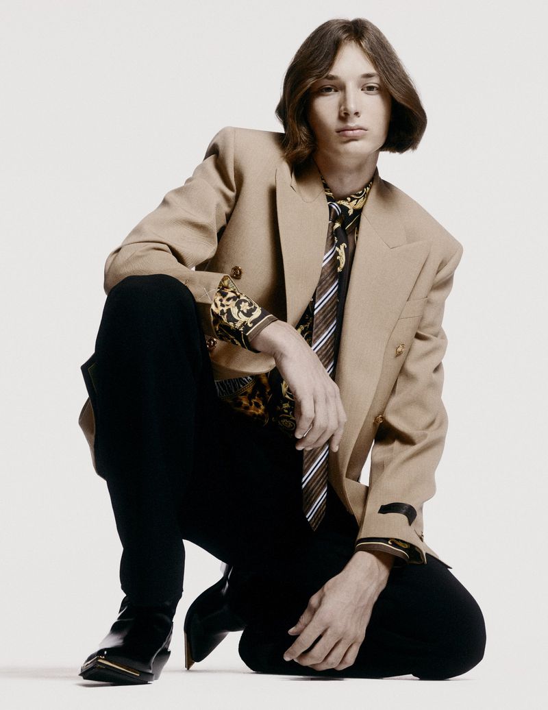 Man in tan blazer, black pants, and patterned shirt kneels in a studio setting, looking at the camera in a fashion-forward pose.