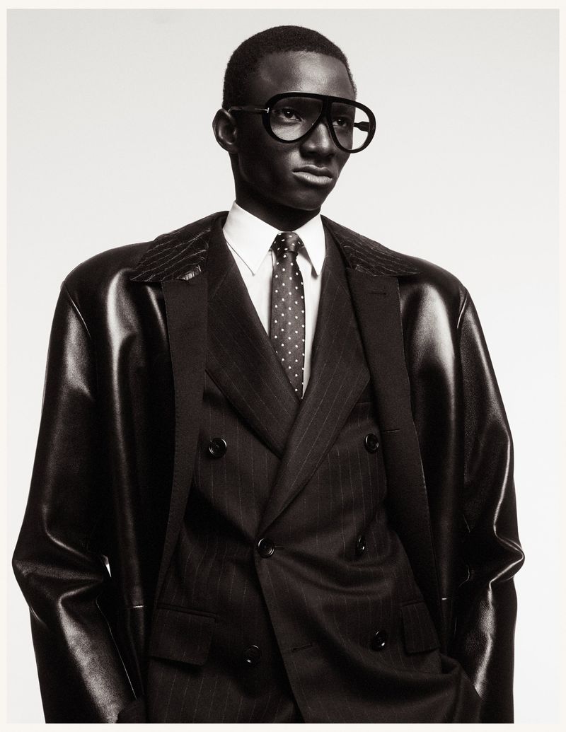 Portrait of a Black man in a pinstripe suit and aviator glasses, posing against a white backdrop in a studio, fashion photography.