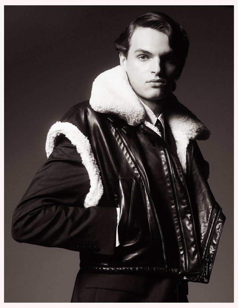 Monochrome portrait of a man in a leather jacket with a white fur collar, a suit jacket, and a tie, against a grey backdrop.
